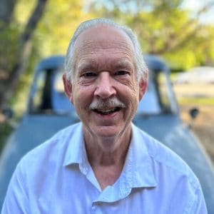 Elderly man smiling outdoors with trees and a vehicle in the background.